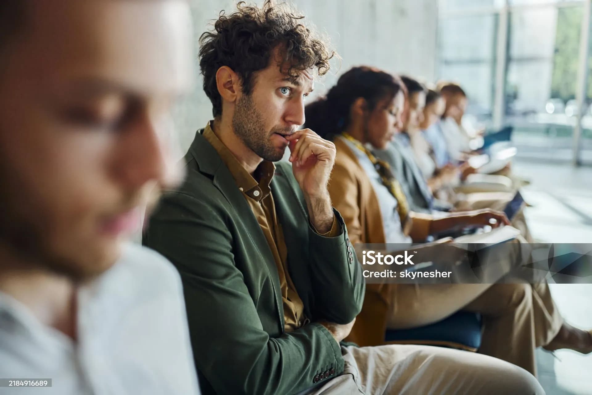 A young man waiting in a row of chairs, hand at his mouth, uneasy.